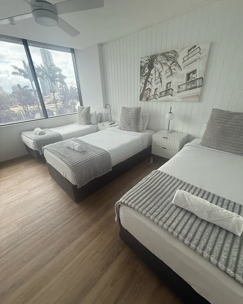 Three single beds made up with white linen, grey throws and rolled towels in a Gold Coast holiday apartment